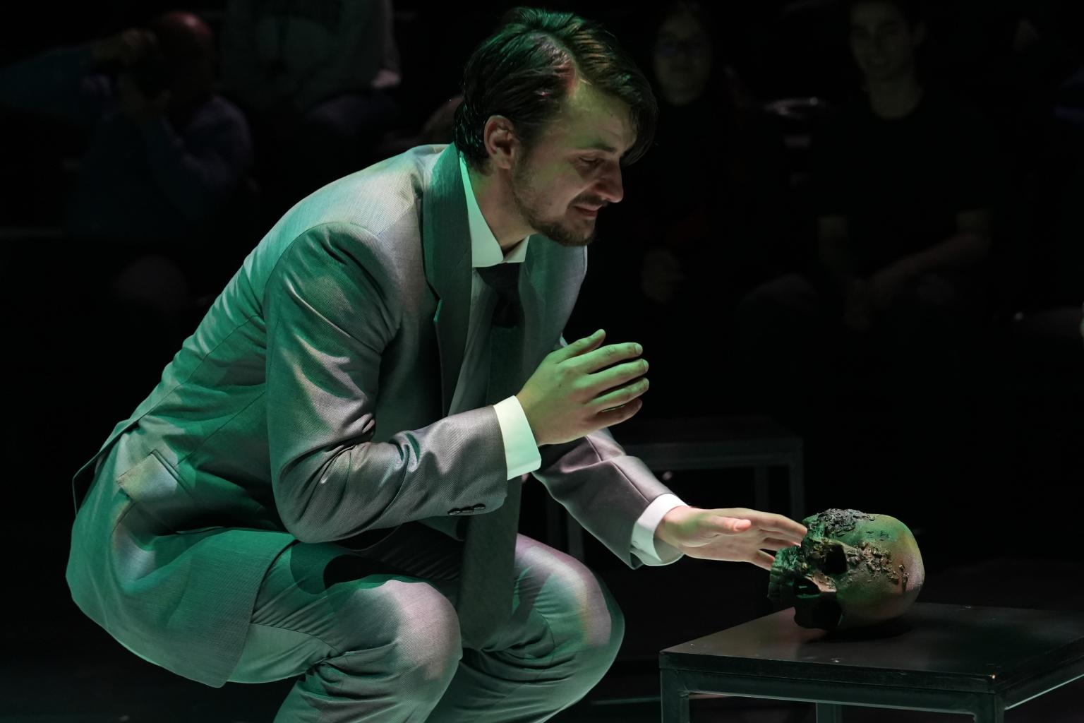 man in grey suit crouched on a dark stage near a skull