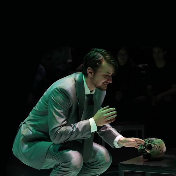 man in grey suit crouched on a dark stage near a skull